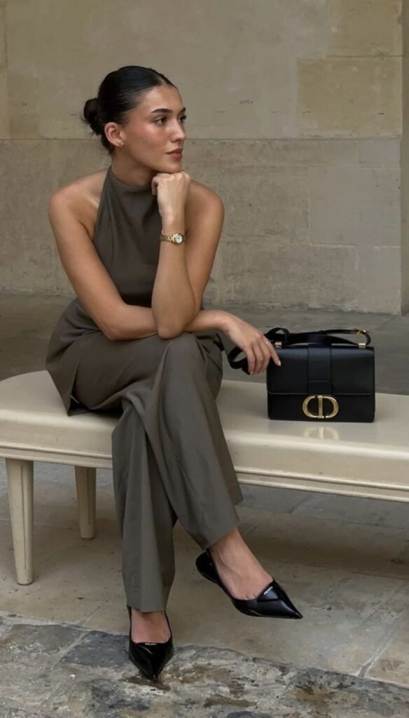 Stylish woman in chic outfit with black handbag, sitting elegantly on a bench indoors.
