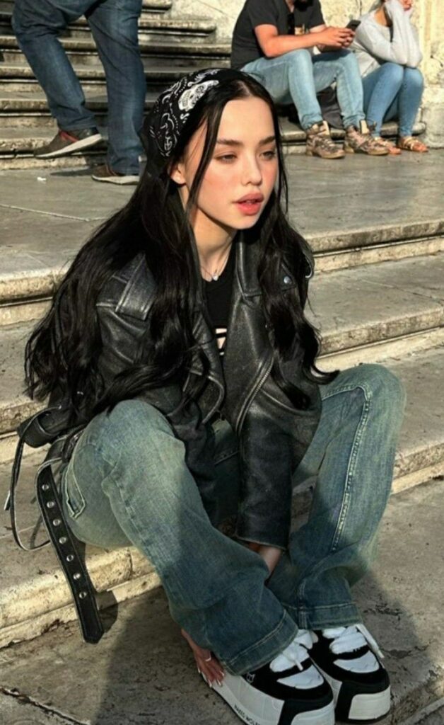 Young woman with long black hair and bandana, wearing jeans and a leather jacket, sitting on stone steps.