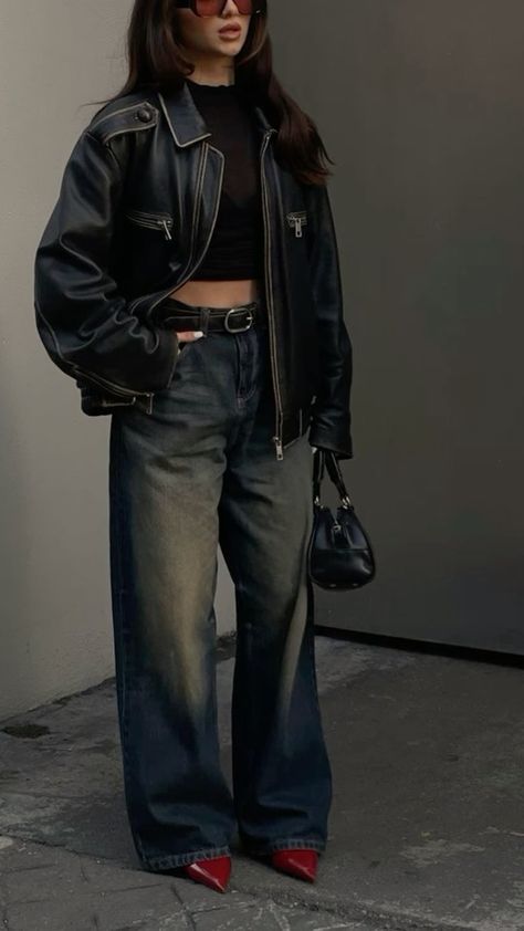 Fashionable woman in black jacket, wide jeans, and red heels, holding a stylish purse. Trendy streetwear outfit.