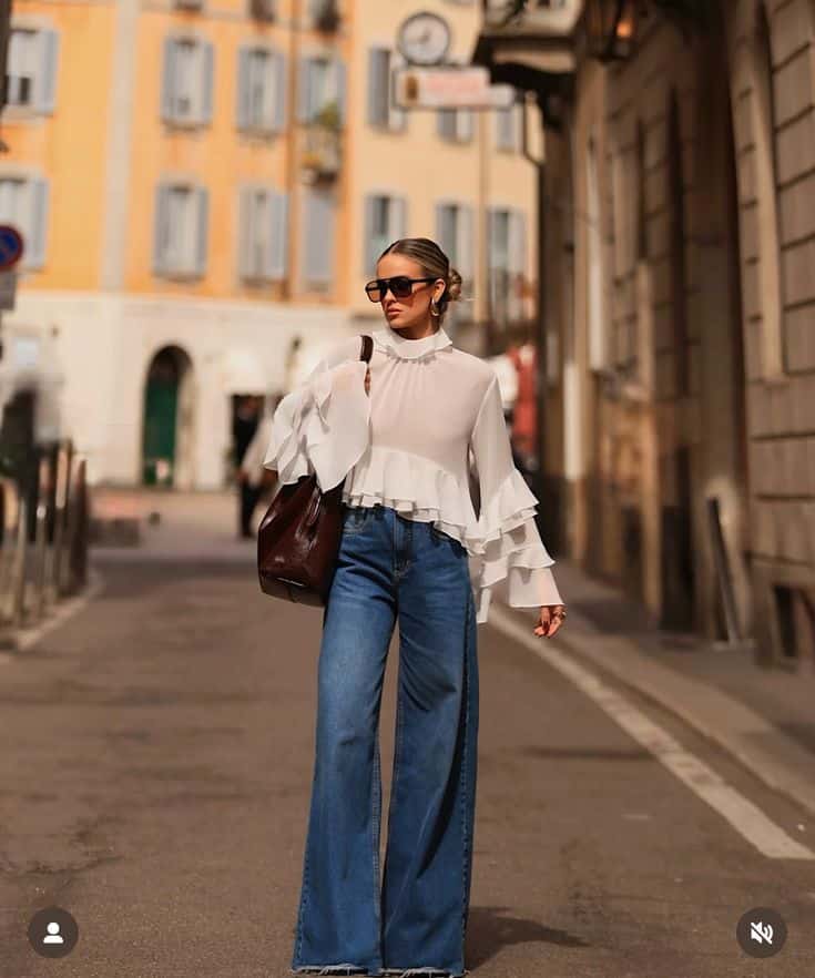 Fashionable woman in sunglasses and white blouse with wide sleeves walks on a stylish European street carrying a large bag.