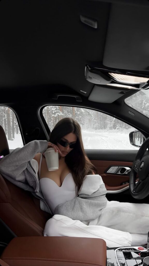 Woman in a car with a white cup, wearing sunglasses and a grey hoodie, snowy forest visible outside the window.