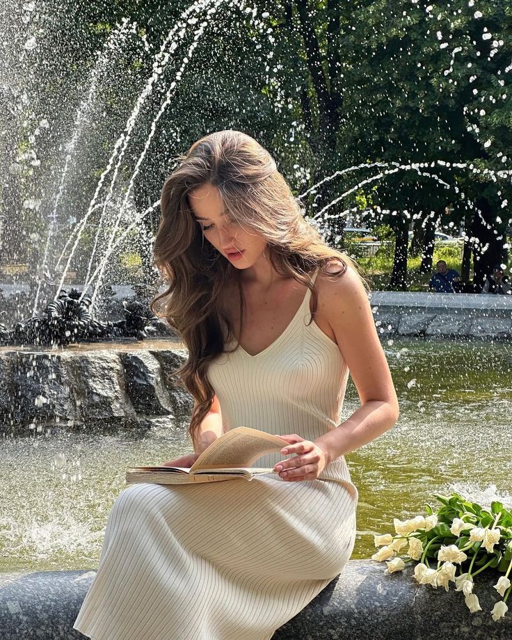 Woman reading by a fountain in a park, wearing a white dress, surrounded by flowers and water.