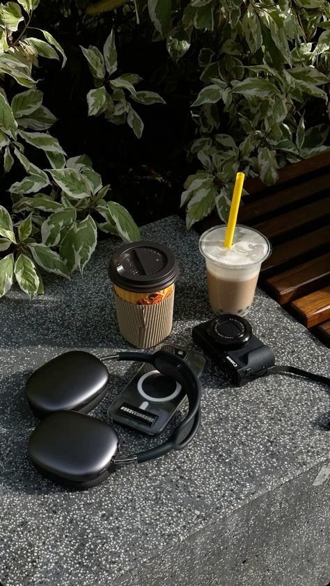 Headphones, camera, coffee, and smoothie on a stone bench with greenery in the background. Optimal outdoor workspace vibes.