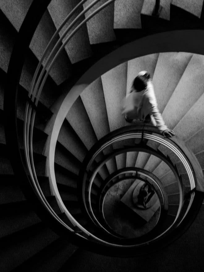 Black and white image of a spiral staircase with a blurred figure descending, creating a dramatic geometric pattern.