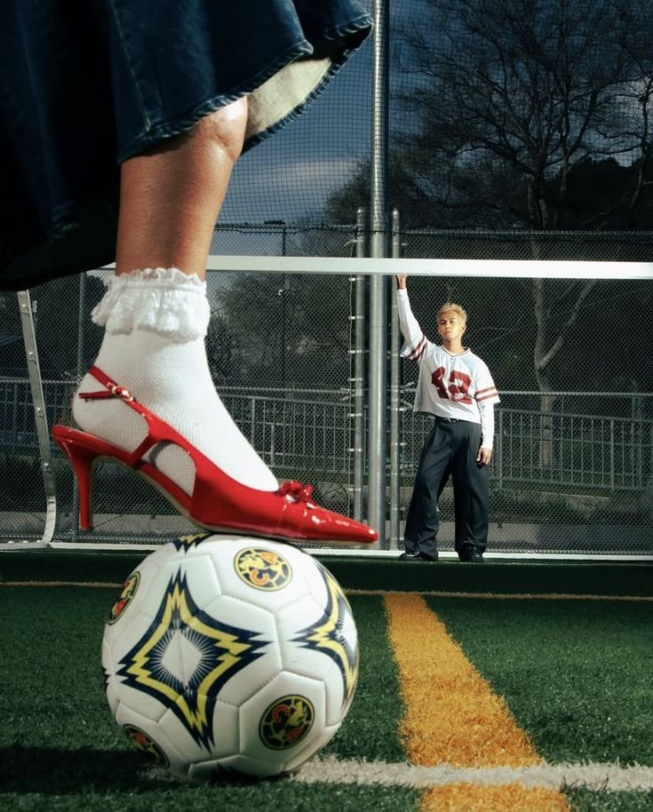 Fashionable red heels on a soccer ball with a player in the background on a sports field.