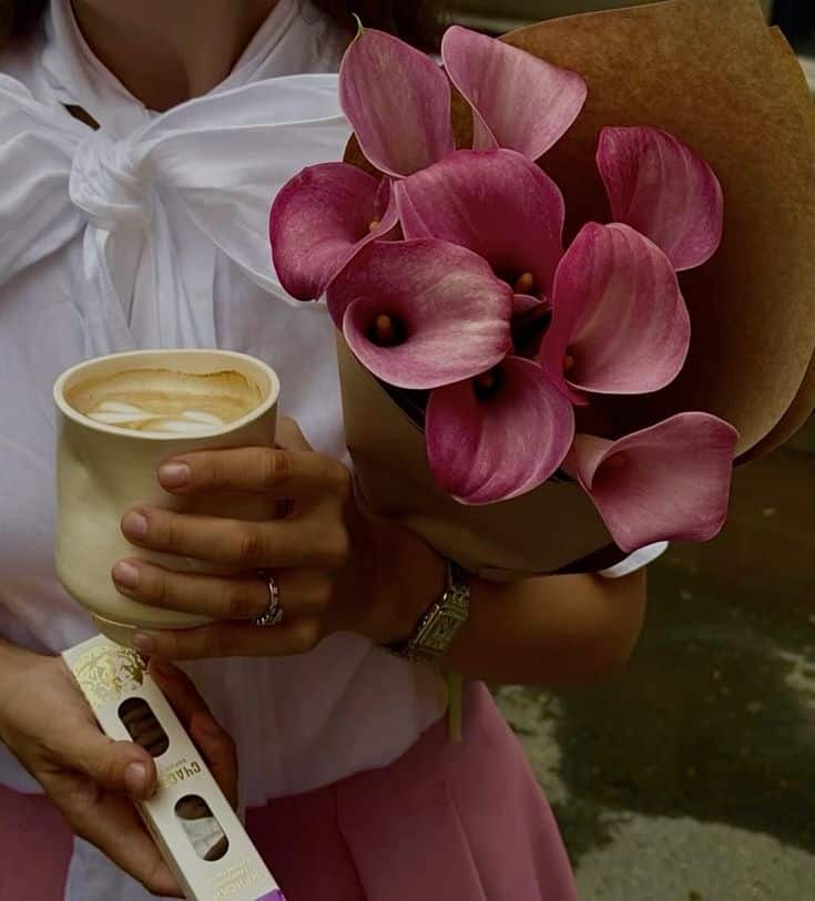 A woman holds a latte, a bouquet of pink calla lilies, and a perfume box, wearing a white blouse and pink skirt.