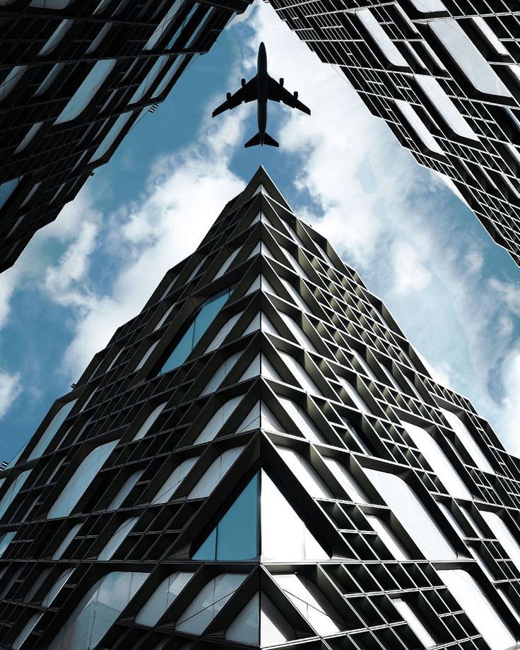 Airplane flying over modern skyscrapers with geometric patterns against a blue sky.