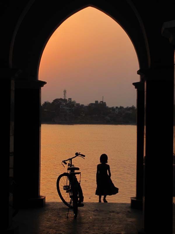 Silhouette of a child and bicycle by a lake at sunset, framed by an archway, creating a serene, scenic view.