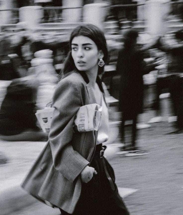 Woman in a checked blazer, holding newspapers, walks through a busy street, monochrome image.