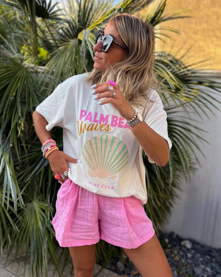 Woman in tropical setting wearing sunglasses, a Palm Beach Waves shirt, and pink shorts, surrounded by palm leaves.