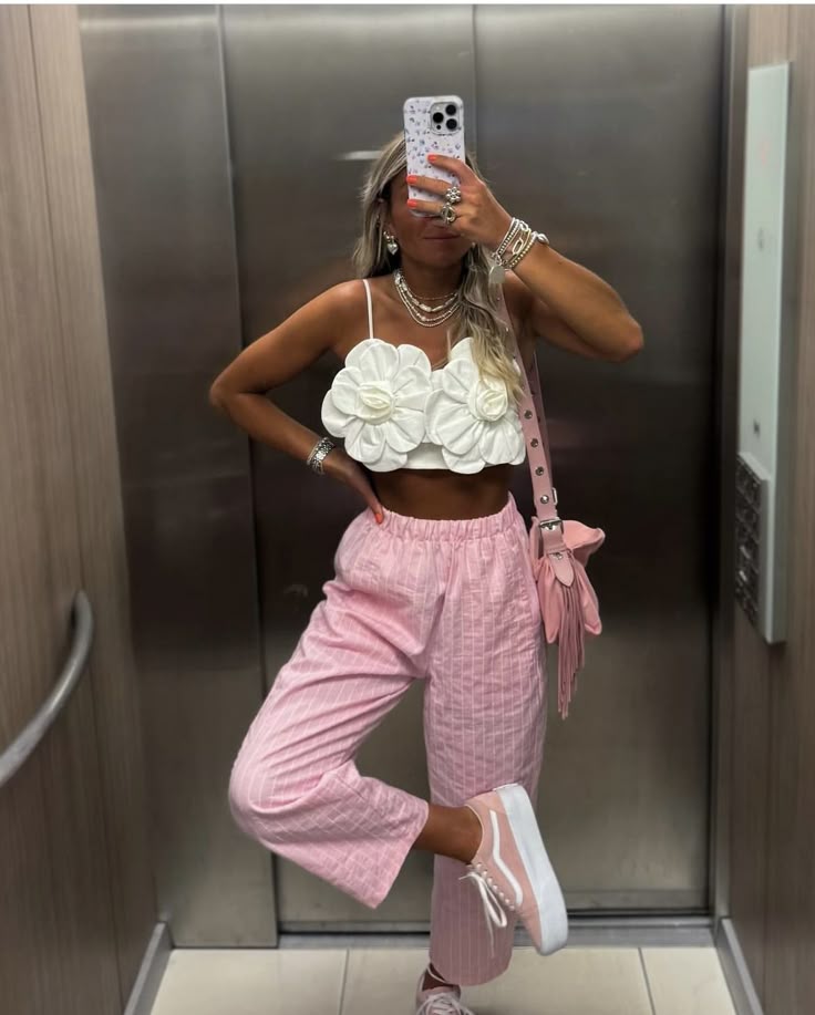 Fashionable woman in an elevator wearing a floral top, pink pants, and sneakers, taking a mirror selfie.