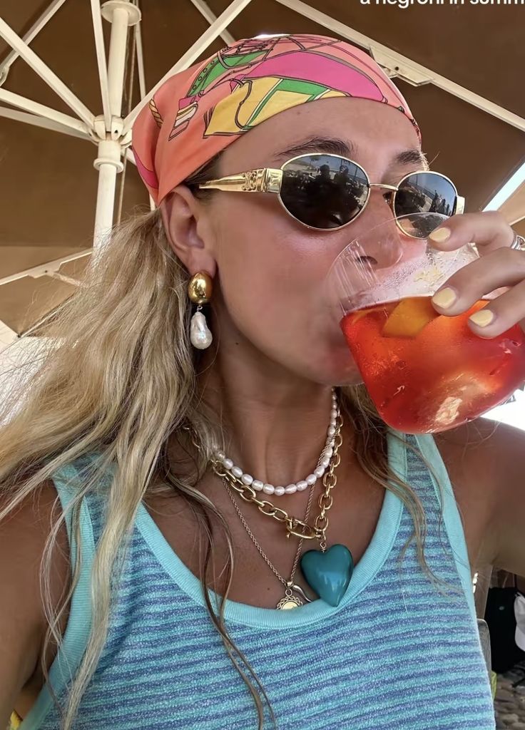 A woman enjoying a refreshing cocktail outdoors, wearing sunglasses and a colorful headscarf, exuding summer style.