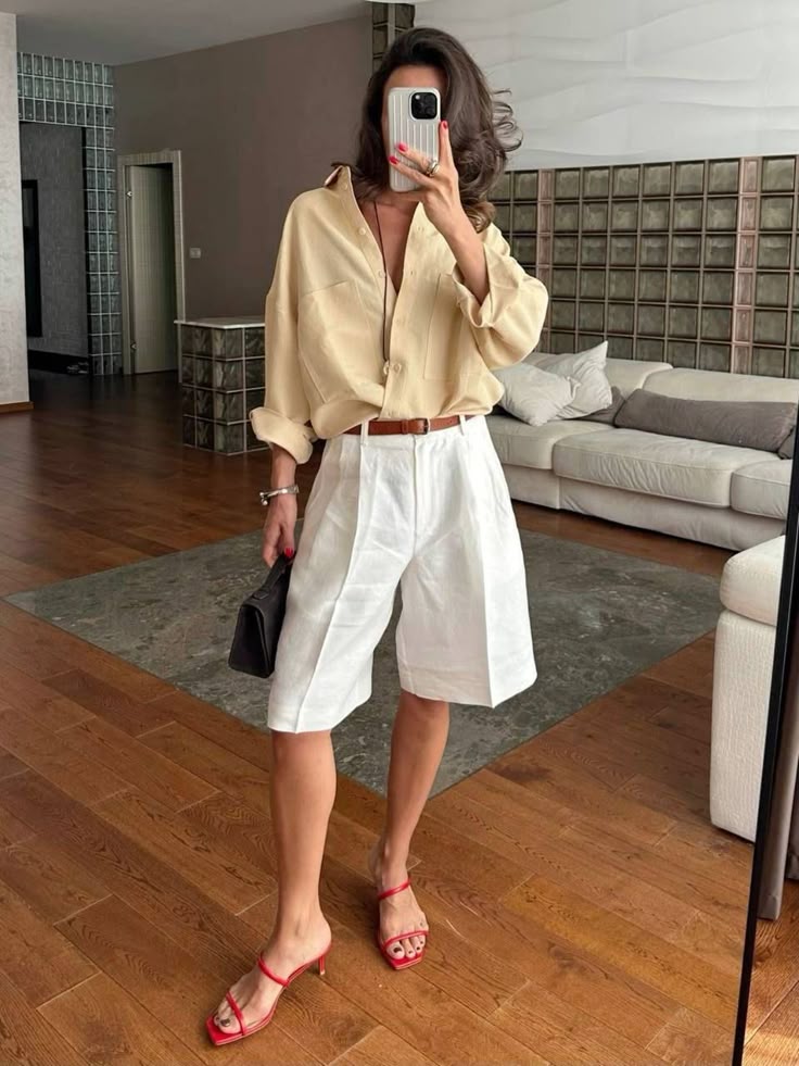 Woman in beige shirt and white shorts takes a mirror selfie in stylish living room.