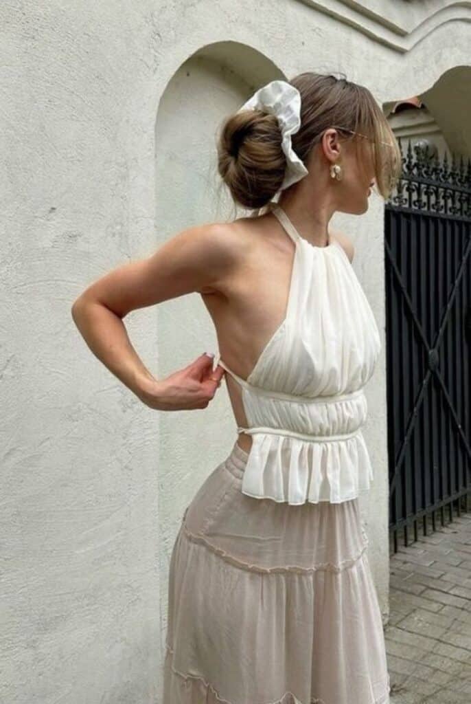 Woman in elegant white halter top and skirt adjusting her outfit outside, with hair in a stylish updo.