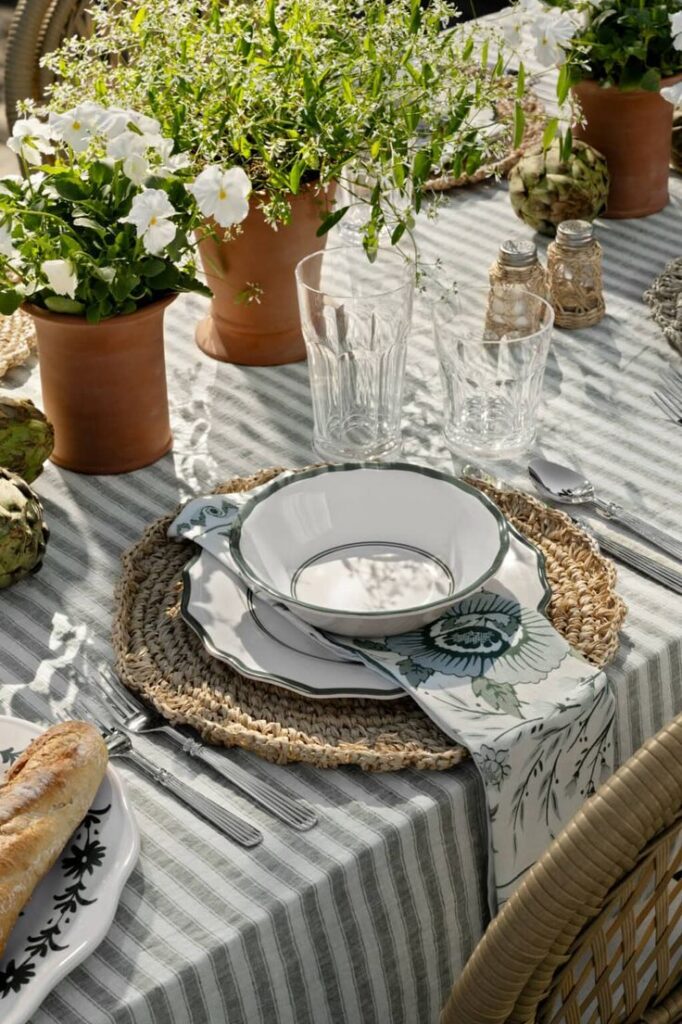 Elegant outdoor dining setup with floral tableware, fresh baguette, and potted plants on a striped tablecloth.