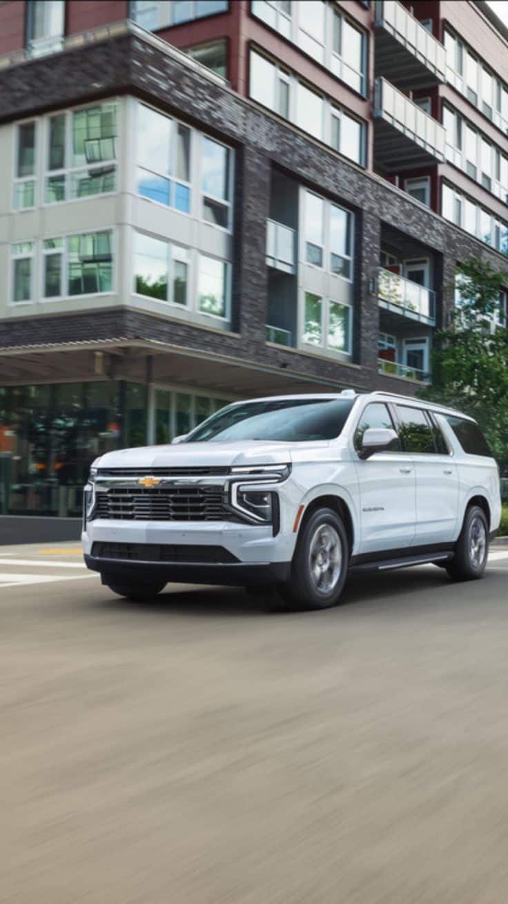 2025 Chevrolet Suburban for Sale _ Spacious Powerful SUV at Gordon Chevrolet White SUV driving past modern apartments in the city. | Sky Rye Design White SUV driving past modern apartments in the city.