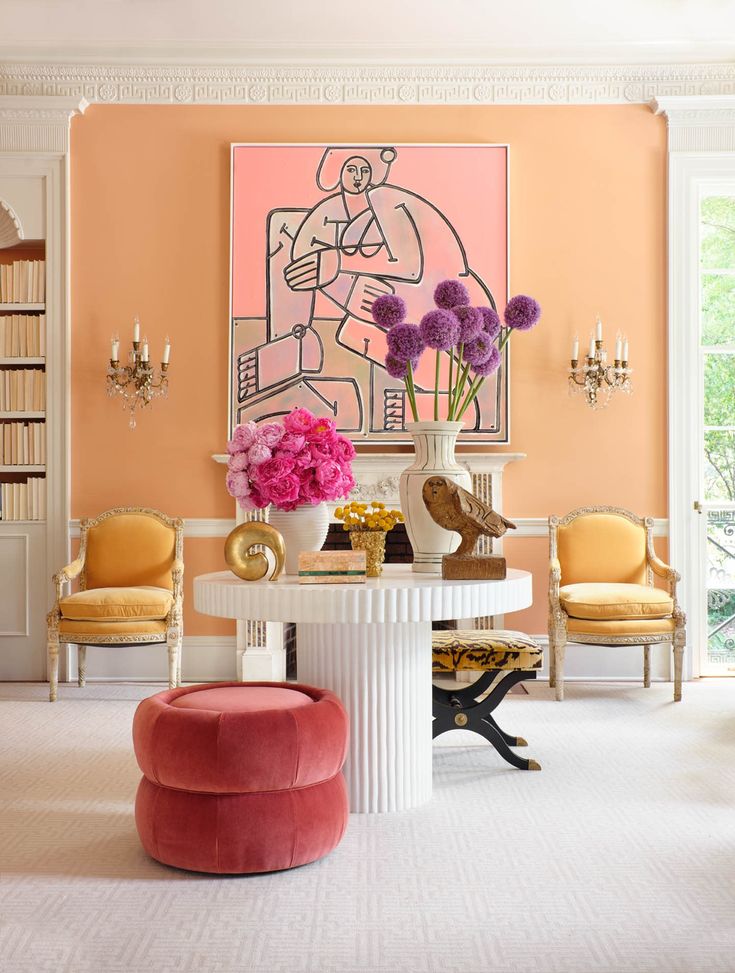Elegant living room with peach walls, vibrant art, and plush pink seating. Decor includes flowers and vintage chairs.