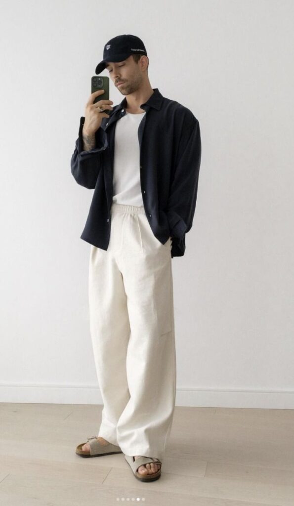 Man in casual style: black cap, navy shirt, white pants, sandals, taking a mirror selfie in a minimalistic room.