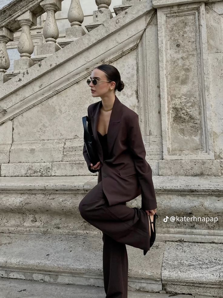 Fashionable woman in a dark suit and sunglasses posing on stone steps, showcasing urban style and elegance.
