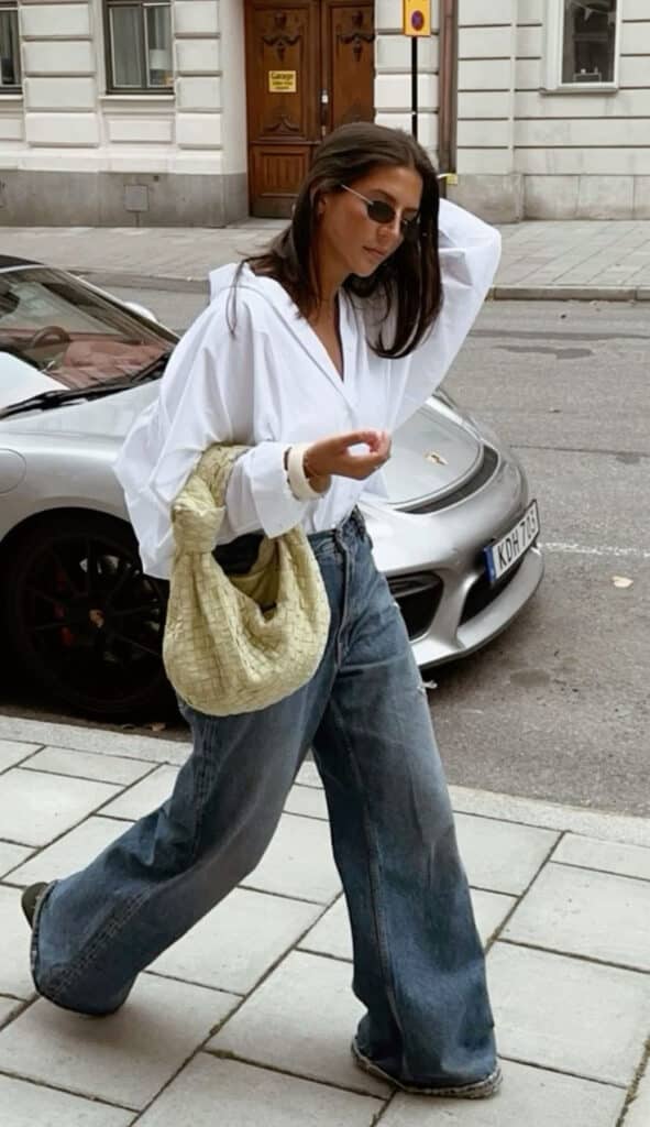 Stylish person in sunglasses and white shirt walking with a woven bag on a city street.