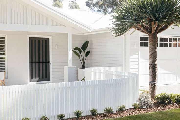 Modern white house exterior with a curved white fence, lush garden, and a tall tree on a sunny day.