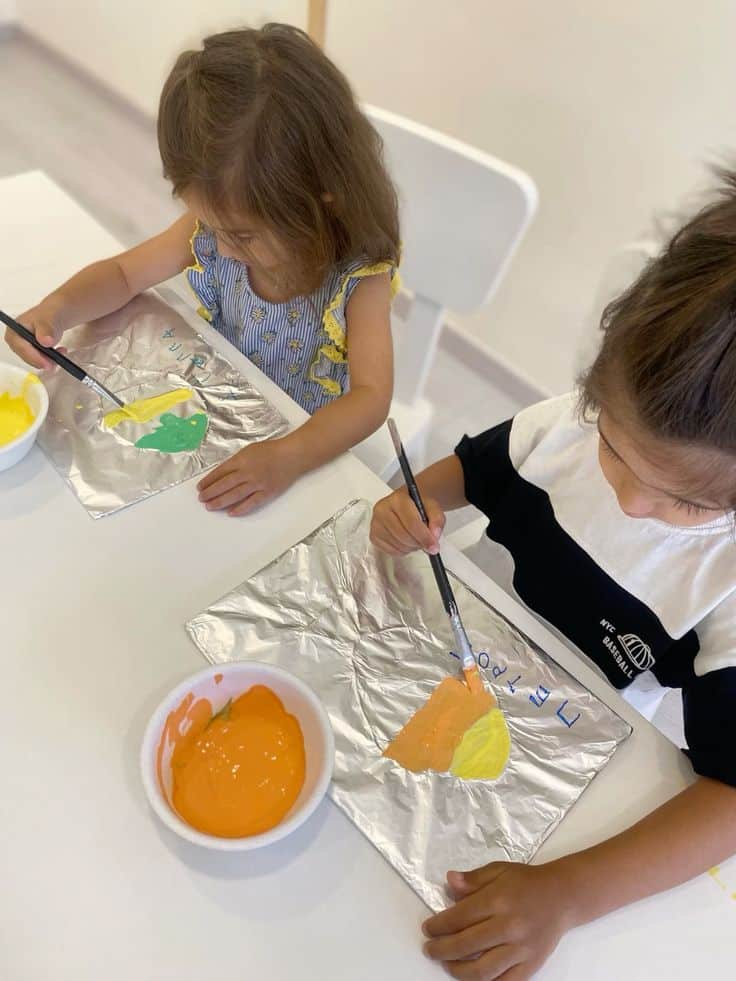 Children painting with brushes and vibrant colors on foil sheets at a table.
