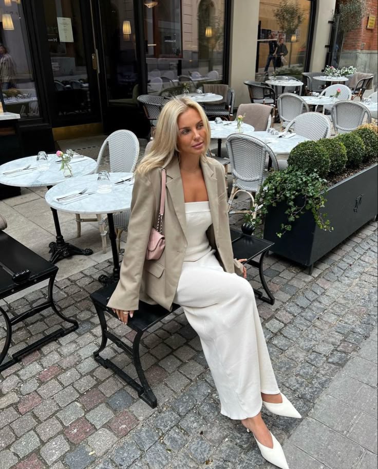 Stylish woman in beige blazer and white dress sitting at a chic outdoor café with cobblestone street ambiance.
