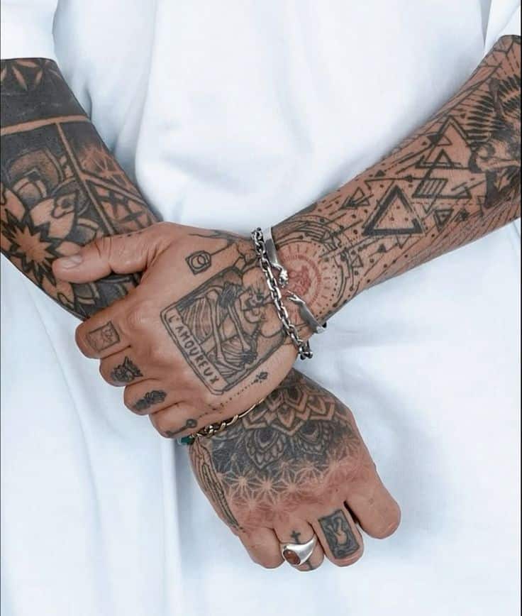 Close-up of intricately tattooed hands and arms with geometric and tribal designs, wearing silver rings and bracelets.