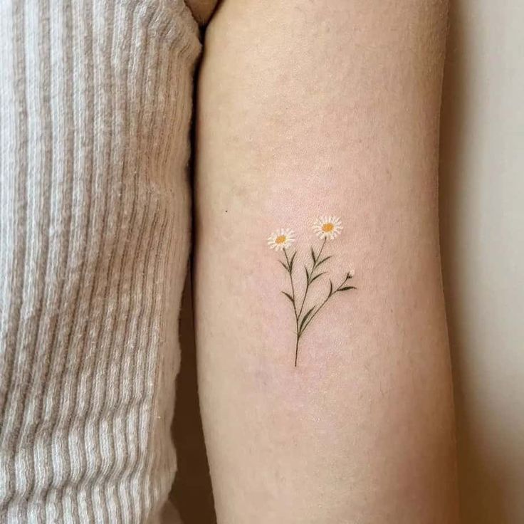 Minimalist daisy tattoo on upper arm, featuring two delicate flowers with green stems.