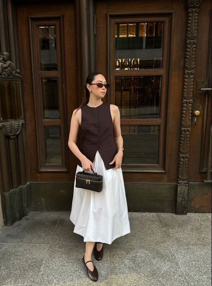 Stylish woman in sunglasses wearing a brown vest and white skirt, holding a purse in front of elegant wooden doors. Summer Office Style Outfit