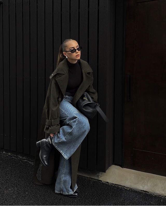 Woman in stylish dark coat and sunglasses poses against black wall, showcasing chic autumn fashion.