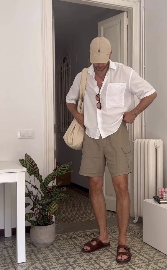 Man in casual summer outfit with shorts, white shirt, and cap stands in a chic, tiled room with a plant.
