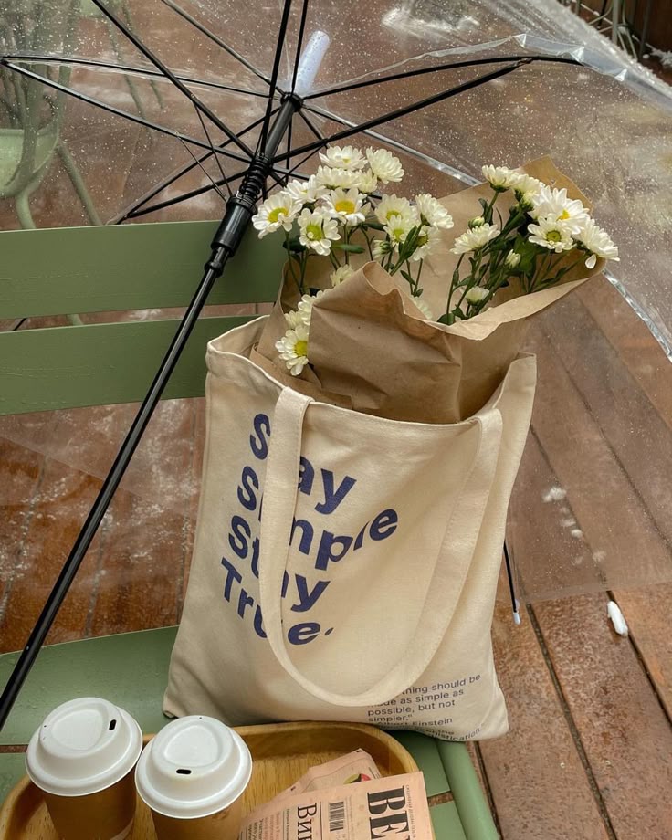 Tote bag with daisies and quote, coffee cups on tray, clear umbrella on rainy cafe table setting.
