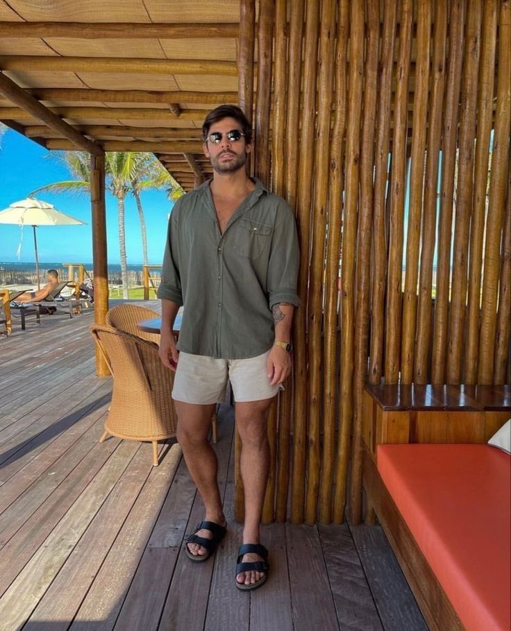 Man in sunglasses and casual summer outfit standing by bamboo wall on a sunny wooden deck with beach view.