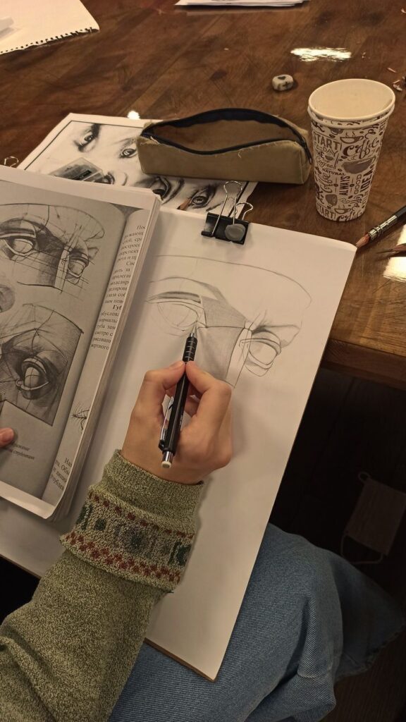 Person sketching an eye with a pencil, using a reference book. Art materials and coffee cup on wooden table.