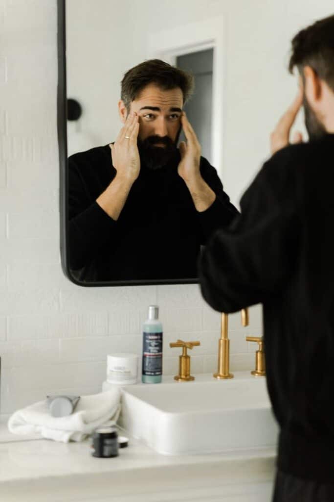 Man applying skincare in front of bathroom mirror with grooming products on sink.
