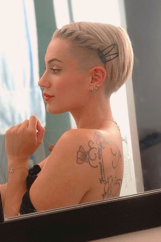 Woman with short blonde hair and braid, tattoos on shoulder, looking in mirror.