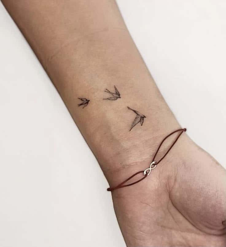 Minimalist bird tattoo on wrist with an infinity bracelet, symbolizing freedom and eternity.