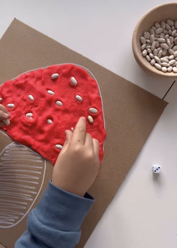 Child creating a mushroom craft with red playdough and white beans on cardboard, enhancing fine motor skills.