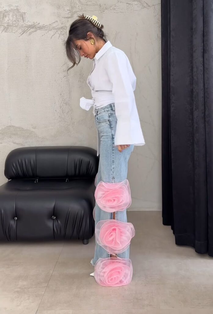 Fashionable woman in jeans with oversized pink floral accents, white blouse, and gold earrings beside a black chair.
