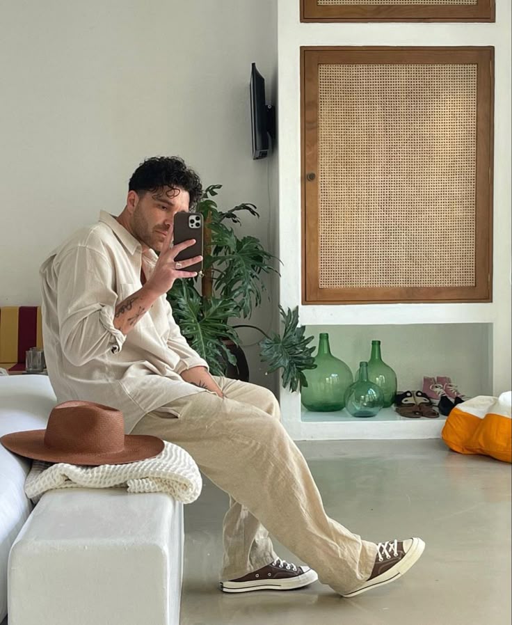 Man in beige outfit taking a selfie in a stylish room with plants and hats, conveying a relaxed, modern vibe. Summer style for men
