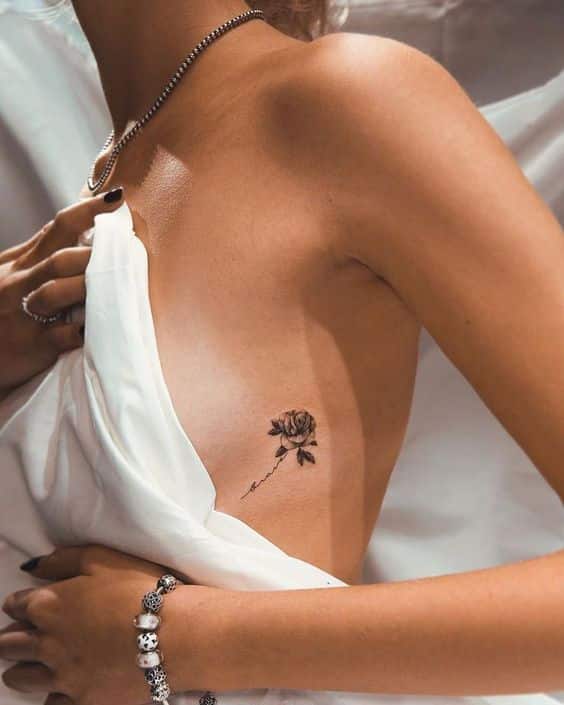 Side tattoo of a rose on a woman's ribcage, partially covered by a white blouse with jewelry visible.