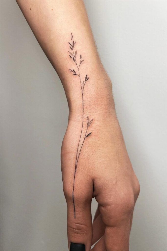 Minimalist plant tattoo on a person's forearm and hand, featuring delicate stems and leaves in black ink.