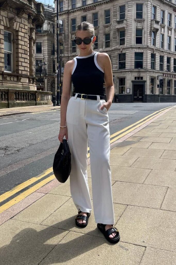 Stylish woman in urban setting wearing white pants, black top, and sunglasses holds a black bag.