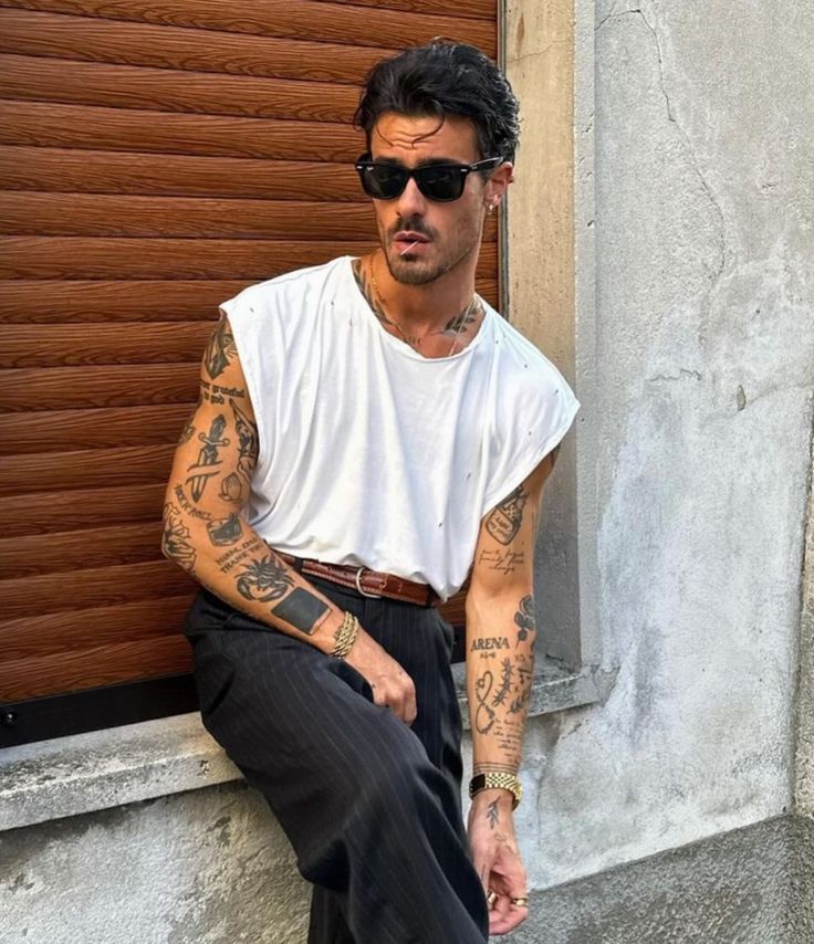 _ 28 Man with sunglasses and tattoos wearing a white sleeveless shirt and striped pants, leaning against a wall. | Sky Rye Design Man with sunglasses and tattoos wearing a white sleeveless shirt and striped pants, leaning against a wall.