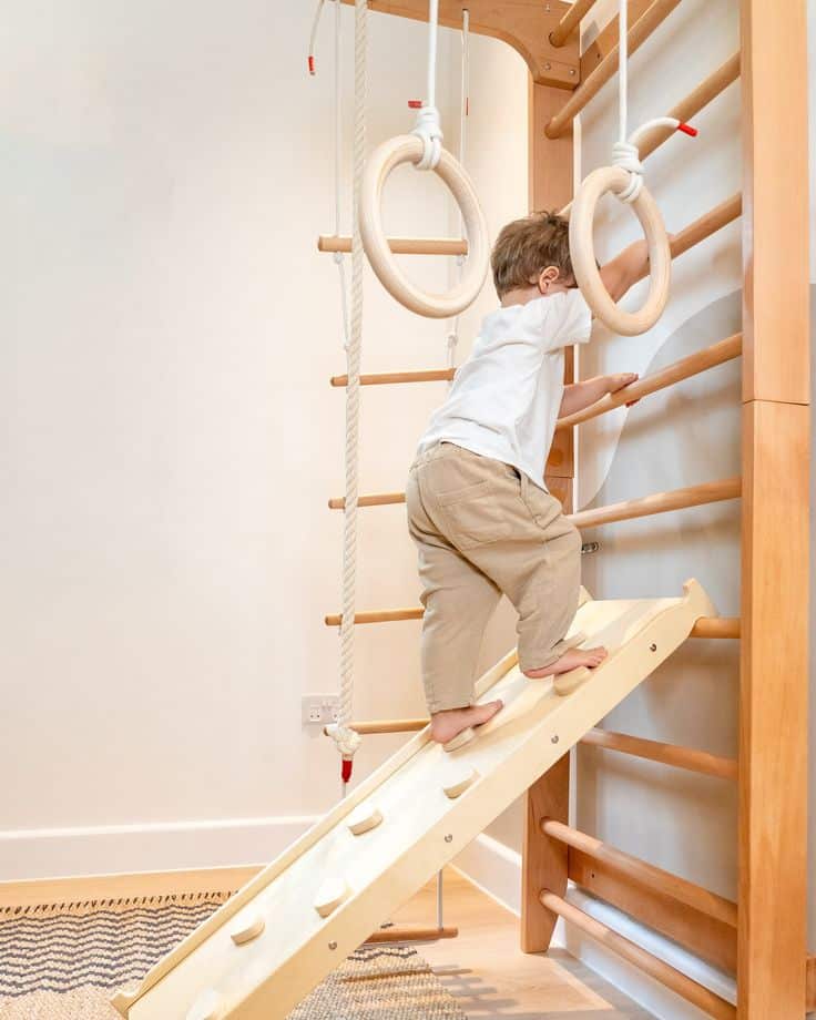 Child climbing indoor play gym with slide, rope, and rings in a bright room for active play.