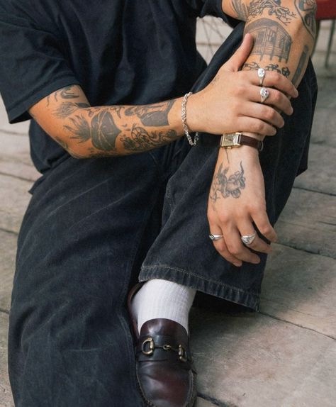 _ 29 Tattooed person wearing black pants, rings, bracelet, and loafers with white socks sitting outdoors. | Sky Rye Design Tattooed person wearing black pants, rings, bracelet, and loafers with white socks sitting outdoors.