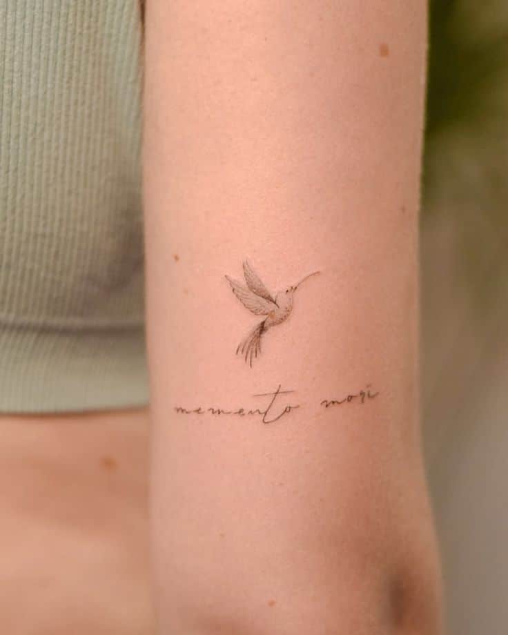 Delicate hummingbird tattoo and memento mori script on upper arm, minimalist design on light skin.