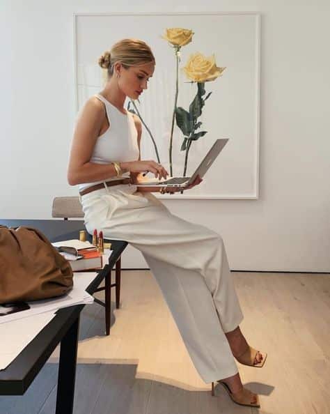 2b67926c-d733-4282-8572-4cb3b030889f Woman in stylish white outfit works on a laptop, sitting on desk, yellow rose artwork in background. | Sky Rye Design Woman in stylish white outfit works on a laptop, sitting on desk, yellow rose artwork in background.