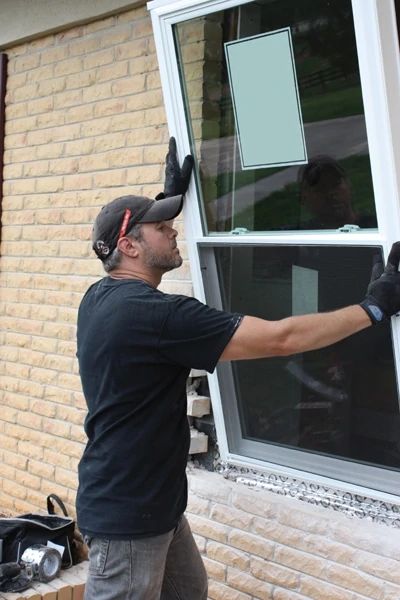 Worker installing a new window on a brick building, wearing gloves and cap for safety.